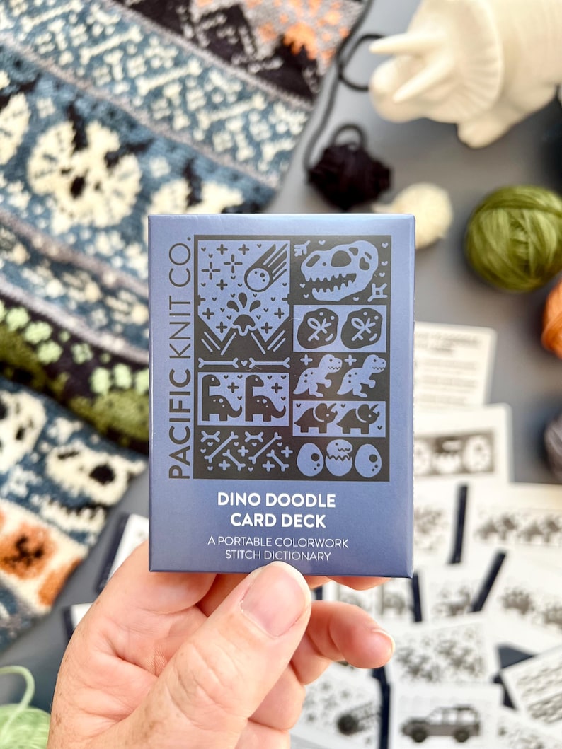 May include: A blue card deck titled "Dino Doodle Card Deck" by Pacific Knit Co. The card deck features dinosaur-themed illustrations. The text on the card reads "A portable colorwork stitch dictionary". The background includes yarn and knitted fabric.