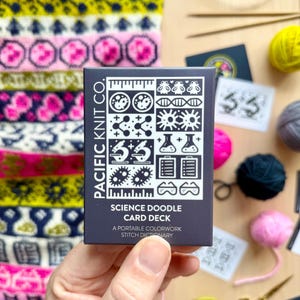 May include: A hand holds a dark blue card deck labeled "SCIENCE DOODLE CARD DECK" with scientific illustrations. Colorful knitted fabric and yarn balls in yellow, pink, and gray surround the deck. Knitting needles and scissors are also visible.