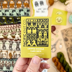 May include: A hand holds a yellow Cryptid Doodle Card Deck by Pacific Knit Co. The card deck features black and white illustrations of various cryptids. Knitting supplies, including yarn and scissors, are in the background.