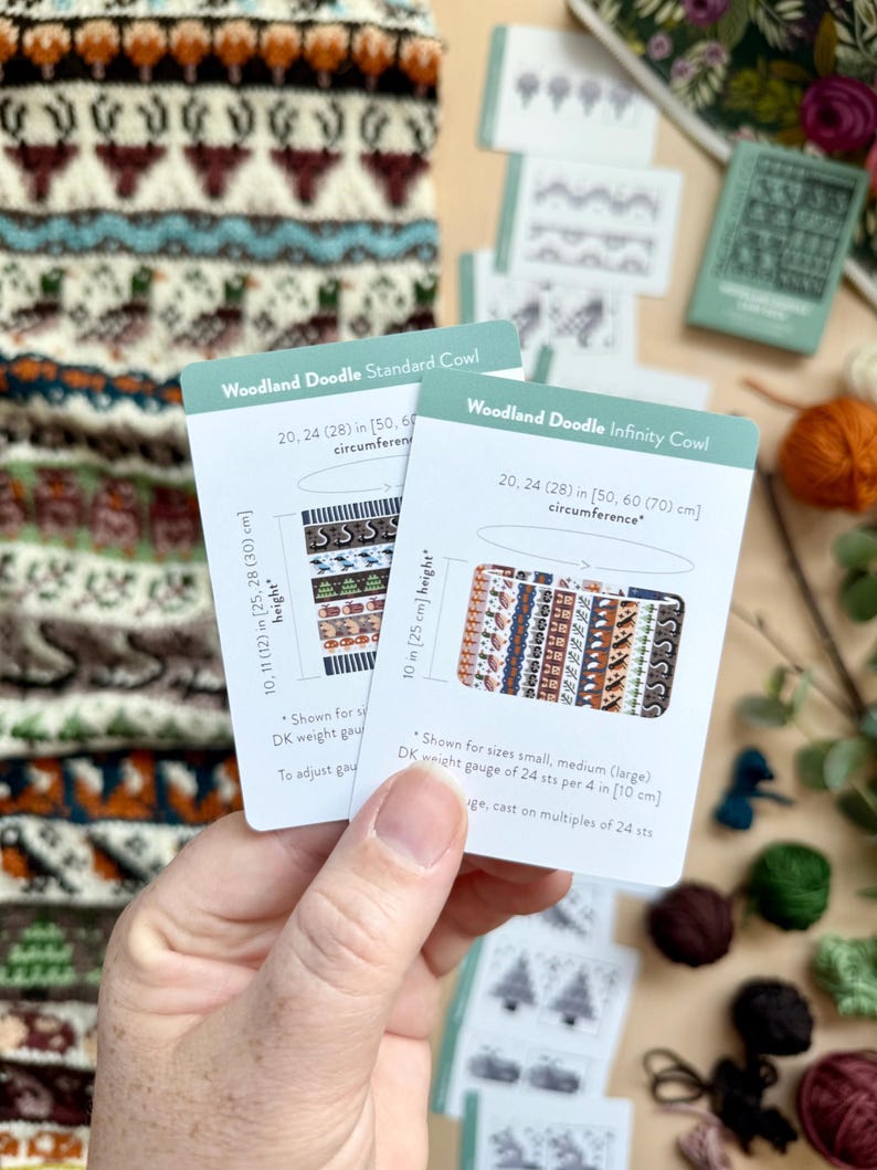 Peut inclure: Deux cartes "Woodland Doodle" sont tenues devant un col tricot&eacute;. Les cartes montrent des motifs et des mesures en pouces et en centim&egrave;tres. Le col pr&eacute;sente un motif r&eacute;p&eacute;t&eacute; de cerfs et de formes g&eacute;om&eacute;triques dans des tons de marron, de sarcelle et d'orange.