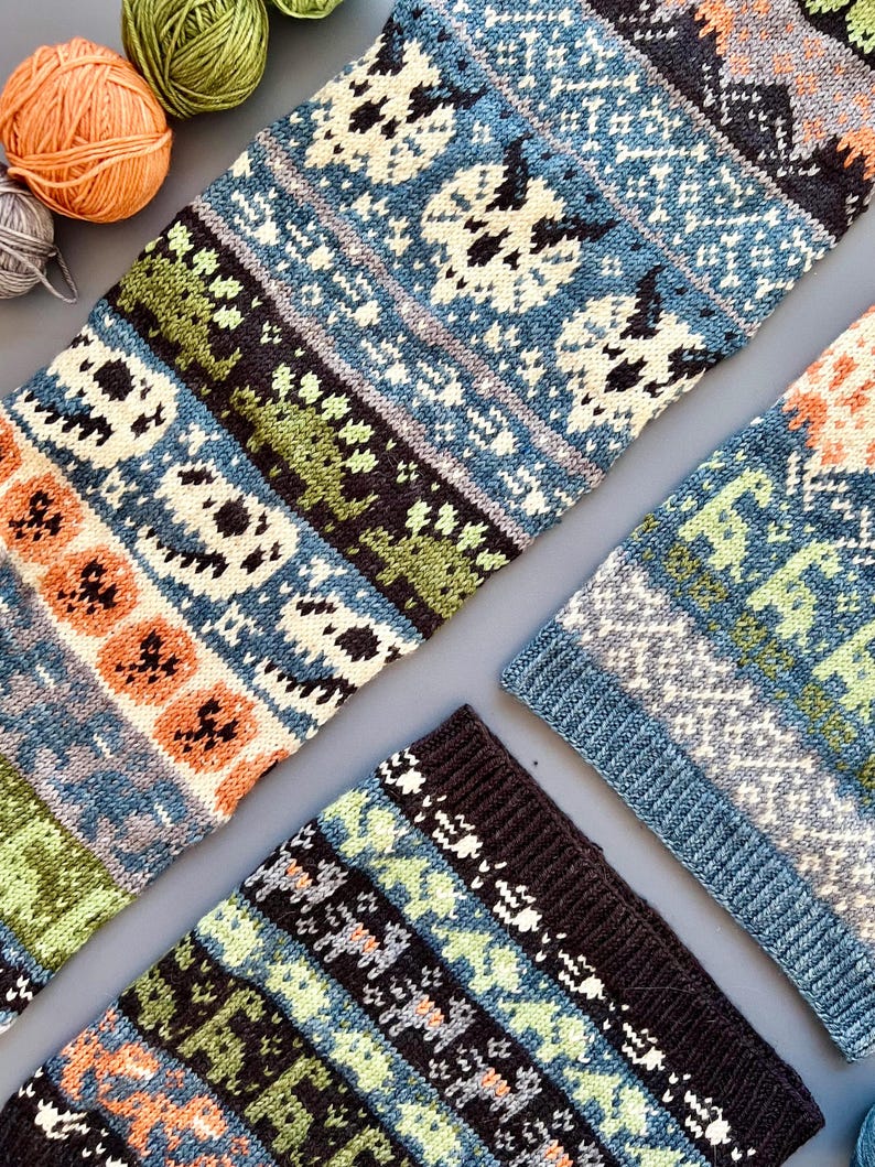 May include: Close-up of several knitted items in shades of blue, green, orange, and black. The items feature repeating patterns of skulls and other geometric shapes. Several balls of yarn in orange, green, and gray are in the upper left corner.