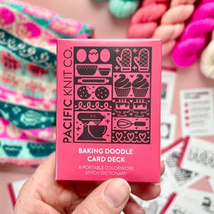Könnte beinhalten: Ein rosa Kartenspiel mit dem Titel "Baking Doodle Card Deck" von Pacific Knit Co. Das Spiel zeigt schwarze Illustrationen von Backwerkzeugen und Leckereien. Der Text "A Portable Colorwork Stitch Dictionary" ist ebenfalls sichtbar. Der Hintergrund umfasst farbenfrohes Garn und Strickzubehör.