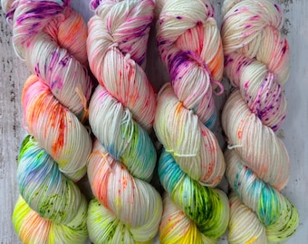 May include: Four skeins of hand-dyed yarn in a vibrant array of colours. The yarn is primarily off-white with splashes of neon yellow, green, orange, pink, and purple. The yarn is arranged on a white wooden surface.