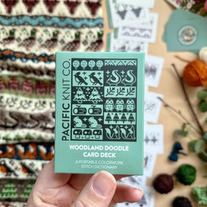 Peut inclure: Une main tient une bo&icirc;te turquoise de cartes "Woodland Doodle Card Deck" de Pacific Knit Co. La bo&icirc;te pr&eacute;sente des illustrations d'animaux des bois. D'autres cartes et un motif tricot&eacute; sont en arri&egrave;re-plan, avec de la laine et de la verdure.