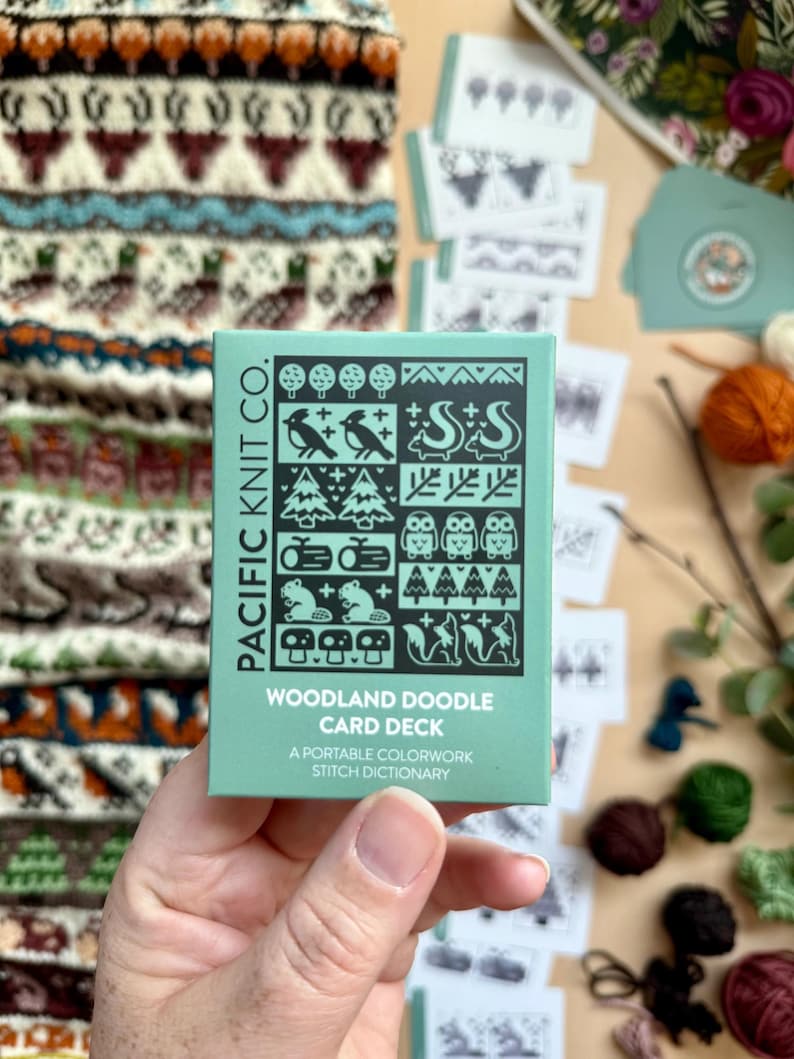 Peut inclure: Une main tient une bo&icirc;te turquoise de cartes "Woodland Doodle Card Deck" de Pacific Knit Co. La bo&icirc;te pr&eacute;sente des illustrations d'animaux des bois. D'autres cartes et un motif tricot&eacute; sont en arri&egrave;re-plan, avec de la laine et de la verdure.