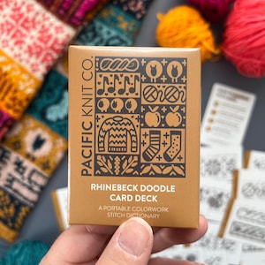 Op de afbeelding: Een bruin kaartspel met de tekst "PACIFIC KNIT CO." en "RHINEBECK DOODLE CARD DECK" in het zwart. Het kaartspel bevat verschillende zwarte illustraties. Kleurrijke bollen wol en gebreide stof zijn op de achtergrond.
