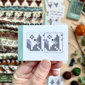 Peut inclure: Une main tient une carte avec un motif de renard pix&eacute;lis&eacute;, intitul&eacute; "Woodland Fox" et "12 sts". La carte pr&eacute;sente un motif de tricot en nuances de gris. D'autres articles li&eacute;s au tricot, y compris du fil et des cartes de motifs, sont &eacute;parpill&eacute;s.