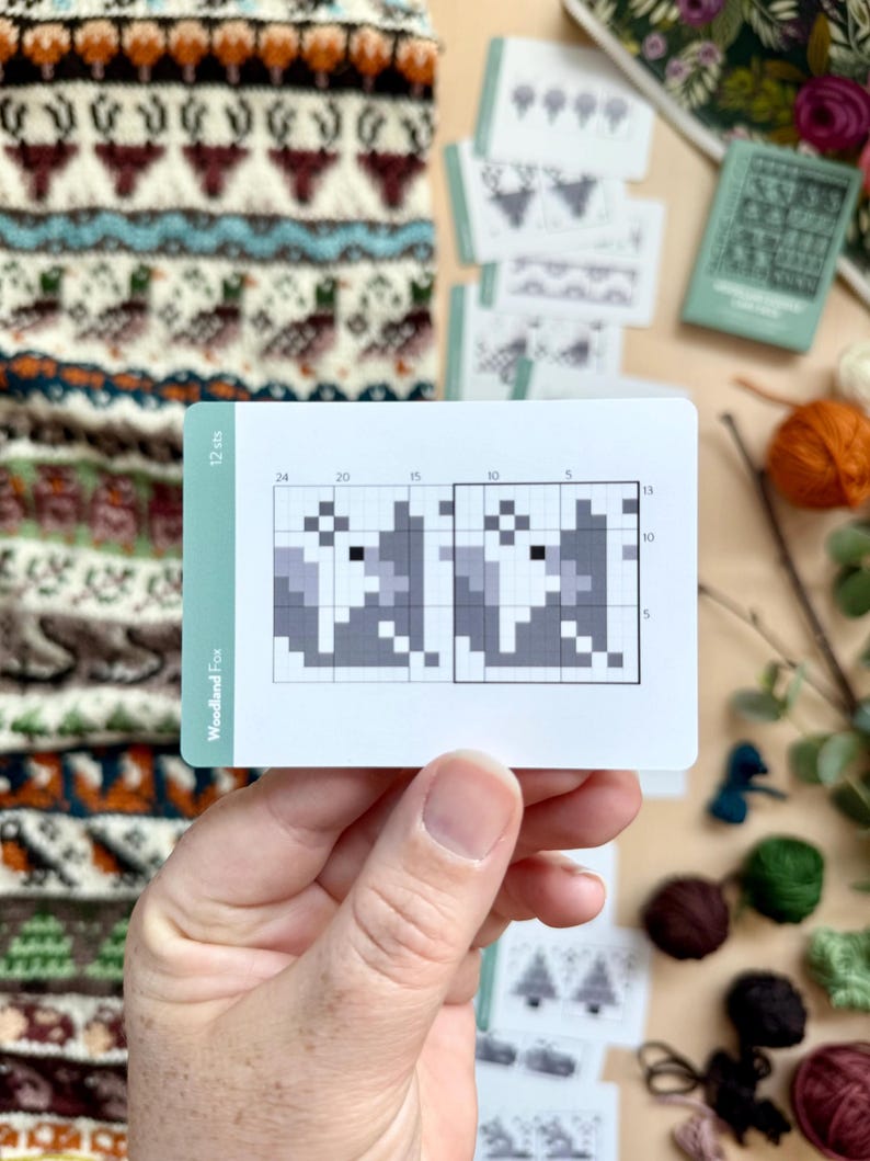 Peut inclure: Une main tient une carte avec un motif de renard pix&eacute;lis&eacute;, intitul&eacute; "Woodland Fox" et "12 sts". La carte pr&eacute;sente un motif de tricot en nuances de gris. D'autres articles li&eacute;s au tricot, y compris du fil et des cartes de motifs, sont &eacute;parpill&eacute;s.