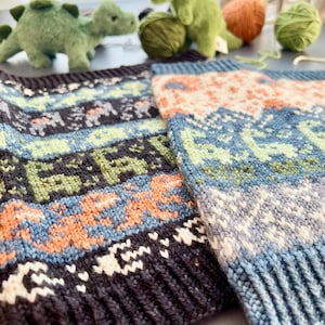 May include: Close-up of two knitted fabric pieces with a colorful pattern. The design features blue, green, orange, and white geometric shapes. The fabric has a ribbed edge. In the background are green dinosaur plush toys and yarn.