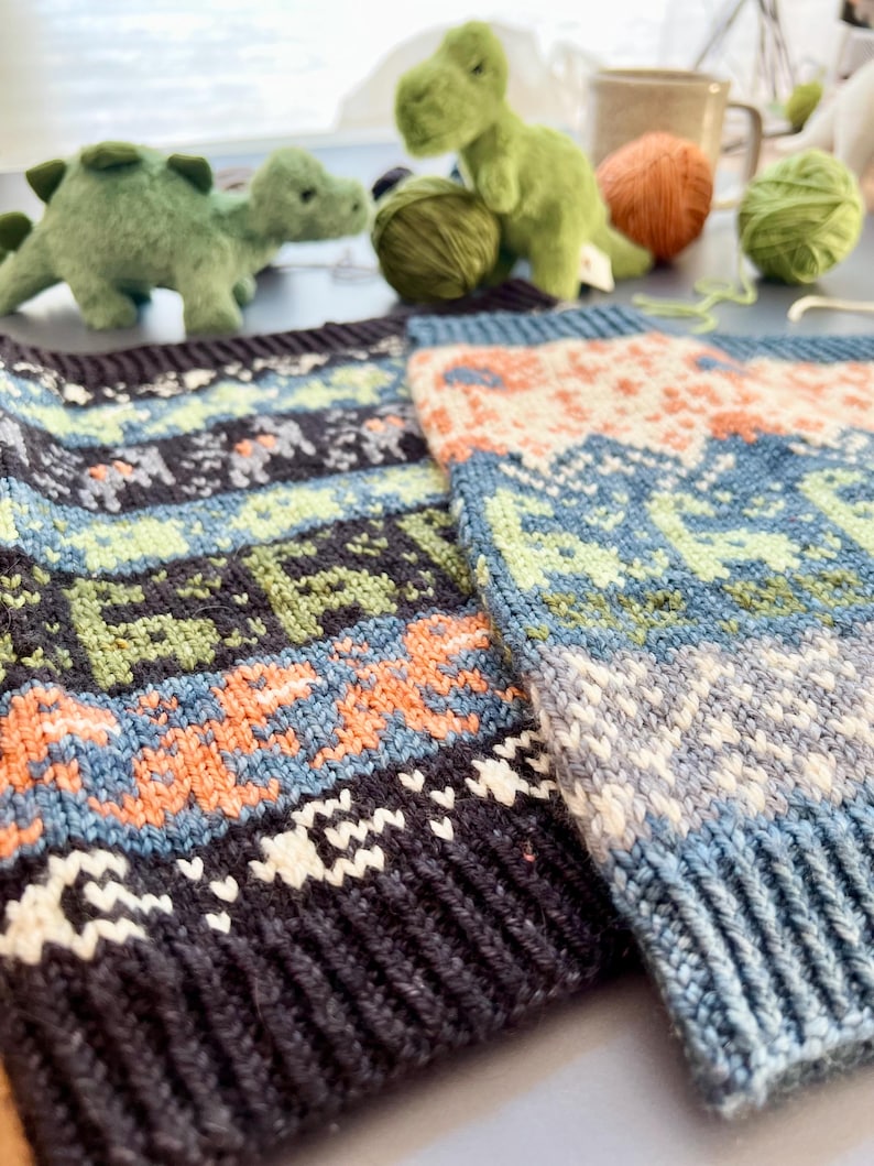 May include: Close-up of two knitted fabric pieces with a colorful pattern. The design features blue, green, orange, and white geometric shapes. The fabric has a ribbed edge. In the background are green dinosaur plush toys and yarn.