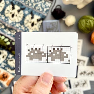 May include: A white card with a pixelated dinosaur design, labeled "Dino Stegosaurus (small)" and "12 sts." The card shows a knitting pattern with a grid and numbers. Various yarn colors and a knitted swatch are in the background.