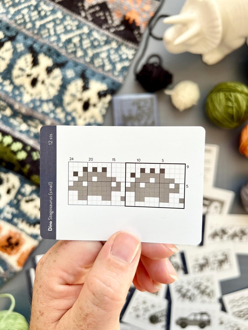 May include: A white card with a pixelated dinosaur design, labeled "Dino Stegosaurus (small)" and "12 sts." The card shows a knitting pattern with a grid and numbers. Various yarn colors and a knitted swatch are in the background.