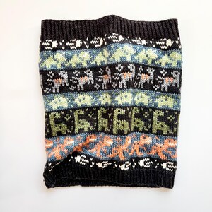 May include: A black knit neck warmer with a colorful geometric pattern. The design includes rows of light blue, green, and orange shapes, along with white and gray animal figures. The neck warmer is made of a soft, textured material.