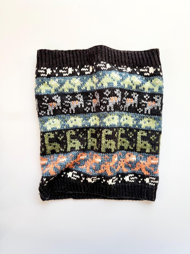 May include: A black knit neck warmer with a colorful geometric pattern. The design includes rows of light blue, green, and orange shapes, along with white and gray animal figures. The neck warmer is made of a soft, textured material.