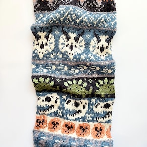 May include: A knitted scarf with a repeating pattern of skulls, pumpkins, and geometric shapes. The scarf features a color palette of blue, green, orange, black, and white. The scarf is folded and appears to be made of wool or a similar material.