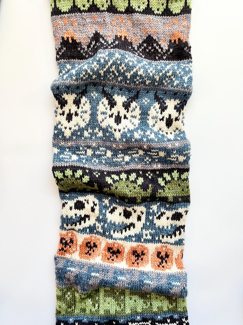 May include: A knitted scarf with a repeating pattern of skulls, pumpkins, and geometric shapes. The scarf features a color palette of blue, green, orange, black, and white. The scarf is folded and appears to be made of wool or a similar material.