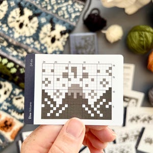 May include: A hand holds a card with a pixelated design labeled "Dino Volcano." The card features a grid with a grayscale pattern resembling a volcano. Knitting supplies, including yarn and a knitted swatch, are visible in the background.