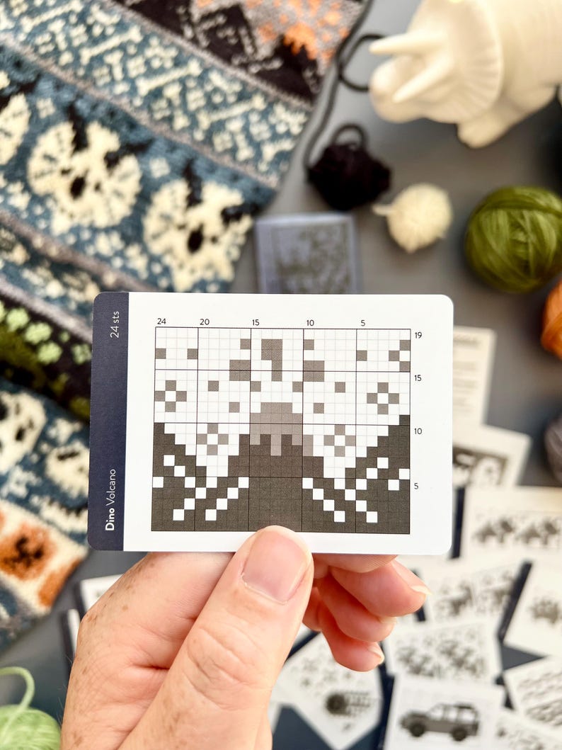 May include: A hand holds a card with a pixelated design labeled "Dino Volcano." The card features a grid with a grayscale pattern resembling a volcano. Knitting supplies, including yarn and a knitted swatch, are visible in the background.