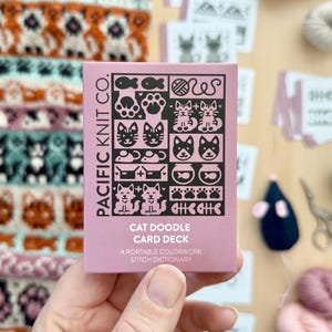 May include: A pink box with the text "PACIFIC KNIT CO." and "CAT DOODLE CARD DECK" on it. The box has a variety of black and white cat-themed illustrations on it. The text "A PORTABLE COLORWORK STITCH DICTIONARY" is also on the box.