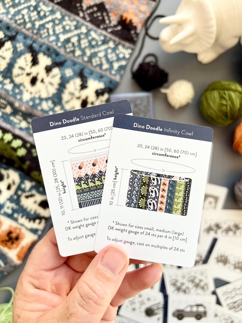 May include: Two cards with "Dino Doodle" cowl patterns are held in a hand. The "Standard Cowl" card shows a colorful design, while the "Infinity Cowl" card displays a pattern with dinosaurs. The cards include measurements in inches and centimeters.