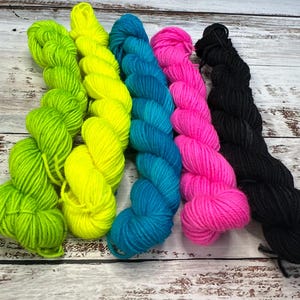May include: Five skeins of colorful yarn in a row. The colors are green, yellow, teal, hot pink, and black. The yarn is arranged on a weathered, light-colored wooden surface.