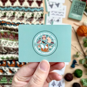 Peut inclure: Une main tient une carte bleu clair avec un logo circulaire indiquant "Doodles by Pacific Knit Co" et "Endless Customization". Le logo pr&eacute;sente des animaux de dessins anim&eacute;s. D'autres cartes et un article tricot&eacute; sont en arri&egrave;re-plan.