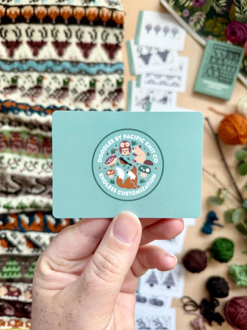 Peut inclure: Une main tient une carte bleu clair avec un logo circulaire indiquant "Doodles by Pacific Knit Co" et "Endless Customization". Le logo pr&eacute;sente des animaux de dessins anim&eacute;s. D'autres cartes et un article tricot&eacute; sont en arri&egrave;re-plan.
