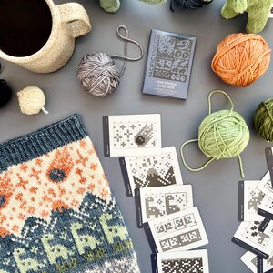 May include: A cozy scene featuring a mug of coffee, yarn balls in various colors, a knitted item with a dinosaur pattern, and a "Dino Doodle Card Deck". Plush dinosaur toys add a playful touch to this craft-themed arrangement.