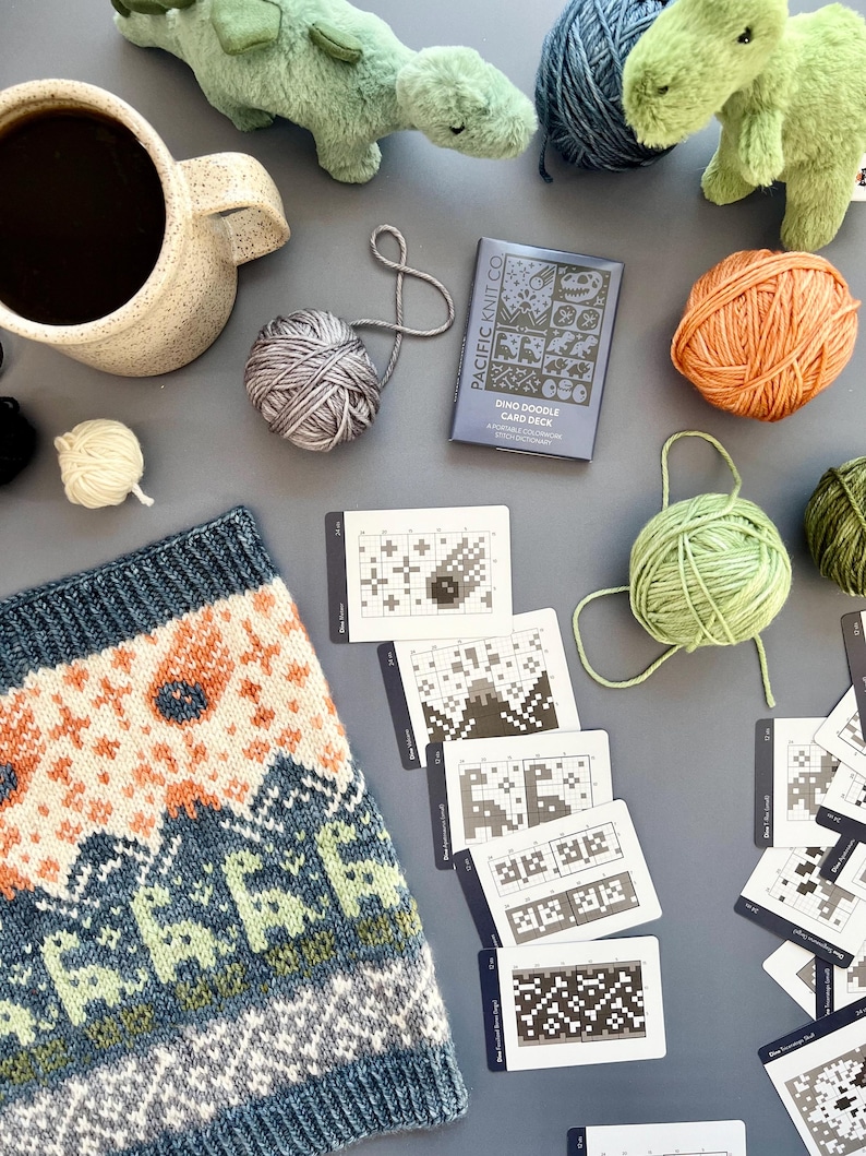 May include: A cozy scene featuring a mug of coffee, yarn balls in various colors, a knitted item with a dinosaur pattern, and a "Dino Doodle Card Deck". Plush dinosaur toys add a playful touch to this craft-themed arrangement.