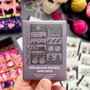 Puede incluir: Una baraja de cartas morada titulada "Spellbound Doodle Card Deck" con varias ilustraciones espeluznantes. El texto de la tarjeta dice "A portable colorwork stitch dictionary." La tarjeta se sostiene en una mano, con suministros de tejido y una calavera en el fondo.