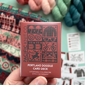 Könnte beinhalten: Eine Hand hält eine rote Kartenspielbox mit schwarzen Illustrationen und dem Text "PORTLAND DOODLE CARD DECK". Die Box ist von bunten Garnknäueln und einem Strickstoff umgeben. Der Text auf der Box lautet ebenfalls "PACIFIC KNIT CO."