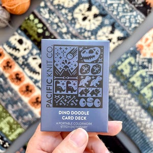 May include: A blue card deck titled "Dino Doodle Card Deck" by Pacific Knit Co. The card deck features dinosaur-themed illustrations. The background includes colorful yarn and knitted patterns with skull and dinosaur designs.