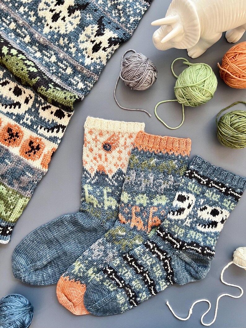 May include: A collection of hand-knitted socks in shades of blue, orange, green, and cream, featuring intricate patterns and designs. The image also includes balls of yarn in various colors and a white ceramic dinosaur figurine, suggesting a craft or hobby theme.