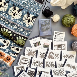 May include: A collection of knitting supplies and patterns. A blue, black, and white knitted piece with a dinosaur design is next to a card deck labeled "Dino Doodle Card Deck" and several yarn balls. The cards show various knitting patterns.