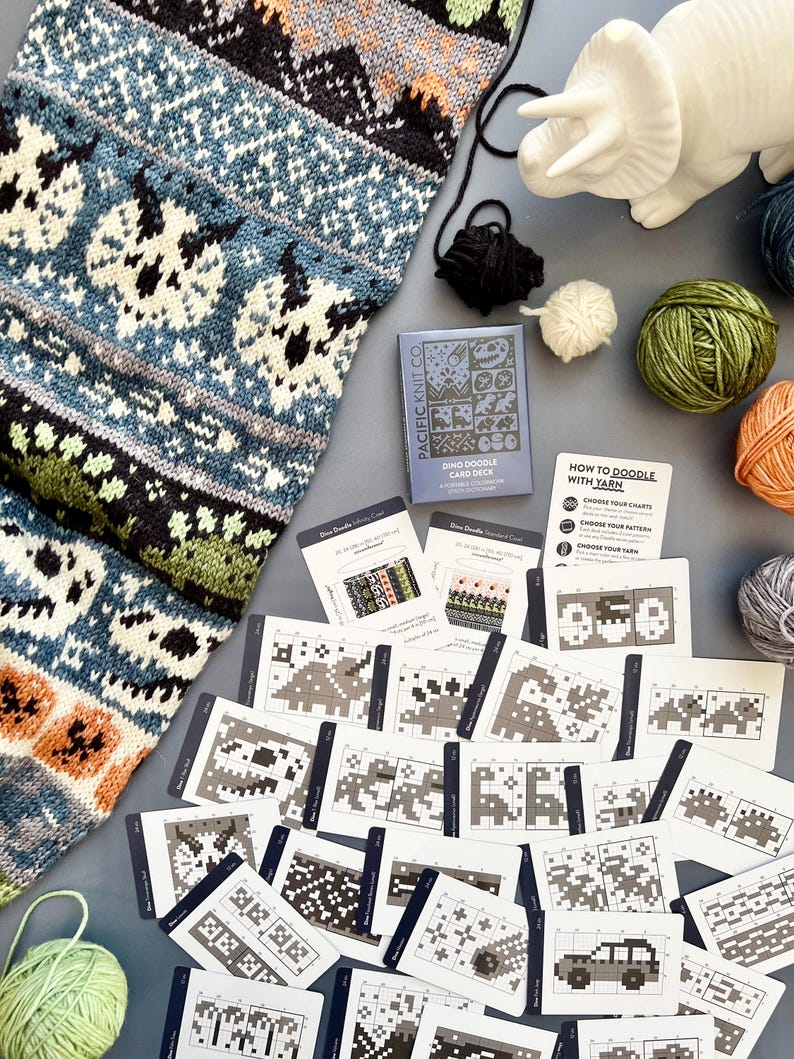 May include: A collection of knitting supplies and patterns. A blue, black, and white knitted piece with a dinosaur design is next to a card deck labeled "Dino Doodle Card Deck" and several yarn balls. The cards show various knitting patterns.