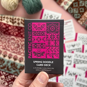 May include: A black card deck held in a hand, featuring a bright pink design with various patterns and the text "SPRING DOODLE CARD DECK". The deck is from "PACIFIC KNIT CO." and is a portable colorwork stitch dictionary. Yarn and knitting samples are in the background.