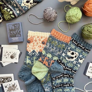 May include: Two pairs of knitted socks with colorful patterns, including dinosaurs and geometric designs. The socks are blue, orange, green, and white. Yarn balls and a card deck with dinosaur illustrations are also visible, suggesting a knitting project.