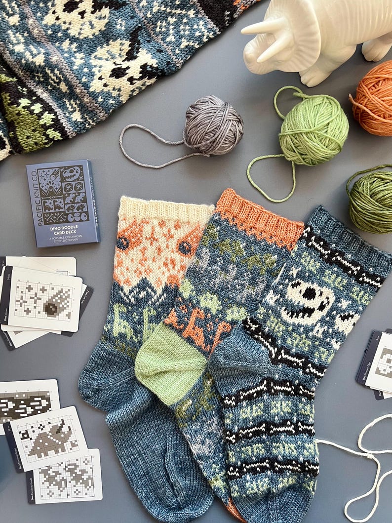 May include: Two pairs of knitted socks with colorful patterns, including dinosaurs and geometric designs. The socks are blue, orange, green, and white. Yarn balls and a card deck with dinosaur illustrations are also visible, suggesting a knitting project.