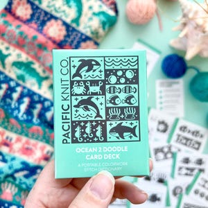 Könnte beinhalten: Ein tealfarbenes Kartenspiel, "Ocean 2 Doodle Card Deck" von Pacific Knit Co, wird in einer Hand gehalten. Das Deck zeigt schwarz-weiße Illustrationen von Meereslebewesen. Ein Strickartikel mit einem Delfinmuster und Wollknäueln befinden sich im Hintergrund.