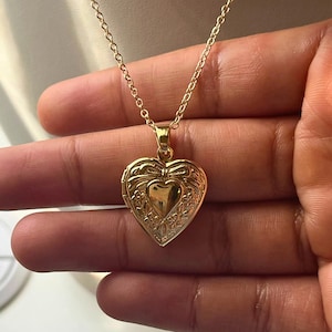 May include: A gold-colored heart-shaped locket necklace. The locket has an ornate design with a bow and floral details. The necklace chain is delicate and also gold-colored. The locket is suspended from the chain.