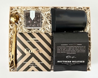 May include: A gift box filled with brown paper shred, containing a black travel mug, a bag of coffee beans, a bag of chocolate, a gold measuring spoon, and a gold clip. The coffee bag has the text "GOODIO CRAFT CHOCOLATE Coffee STONE GROUND IN FINLAND ORGANIC - VEGAN 480 (7 OZ.)" and the text "SOUTHERN WEATHER" on the coffee bag.