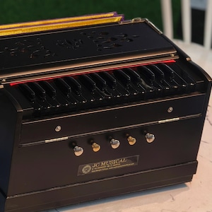 May include: A black harmonium with a keyboard and decorative carvings. The instrument has gold accents and a logo that reads "JC MUSICAL". It sits on a marble-patterned table.