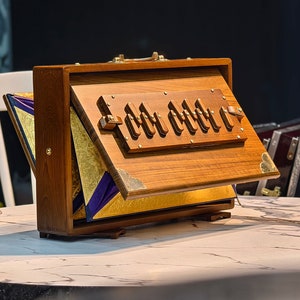 May include: A wooden harmonium with a hinged top and decorative brass hardware. The instrument is open, revealing gold and purple fabric accents. Other harmoniums are visible in the background. The text "JC MUSICAL OFFICIAL" is at the bottom.