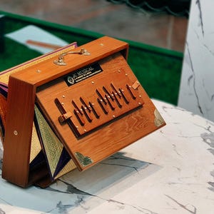 Puede incluir: Un armonio de madera con un asa de latón y detalles decorativos. El instrumento está abierto, revelando las teclas y los componentes internos. El armonio está colocado sobre una mesa con diseño de mármol.