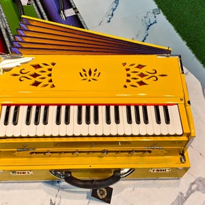 May include: A bright yellow harmonium with a keyboard of white and black keys. The instrument has decorative cutouts and a handle. Other harmoniums in various colors are visible in the background.