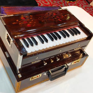 Könnte beinhalten: Ein hölzernes Harmonium mit glänzend dunkelbraunem Finish. Das Instrument hat eine Tastatur mit schwarzen und weißen Tasten, dekorative Schnitzereien und eine ausziehbare Schublade. Das Harmonium steht auf einer weißen Oberfläche.