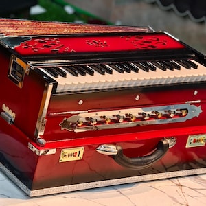 May include: A red and black harmonium with a keyboard and bellows. The instrument has a handle and decorative accents. It sits on a marble-patterned table. The harmonium is a musical instrument.