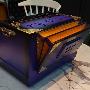 May include: An ornate box with a deep purple exterior and gold detailing. The box features a hinged lid and side panels that open to reveal an orange interior. The design includes cut-out patterns and decorative hardware.