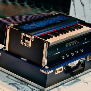 May include: A black and blue harmonium with a keyboard and bellows. The instrument has gold-colored accents and is sitting on a round, marble-patterned surface. The text "JC MUSICAL OFFICIAL" is visible.
