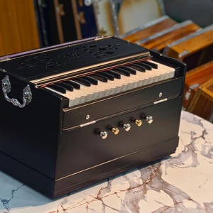 May include: A black harmonium with a keyboard and decorative carvings on top. The instrument has silver handles and knobs. It sits on a marble-patterned table, with other harmoniums in the background.